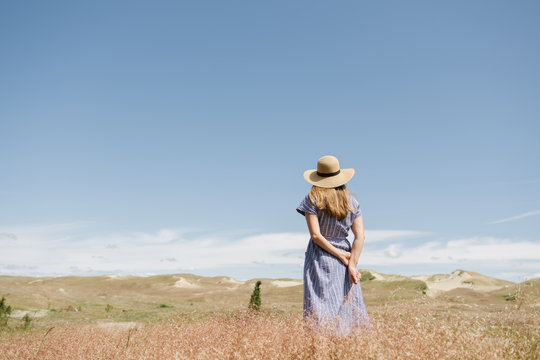 back view of adult woman in dress with wisp of dry grass spikes while standing on scenic field, Nida