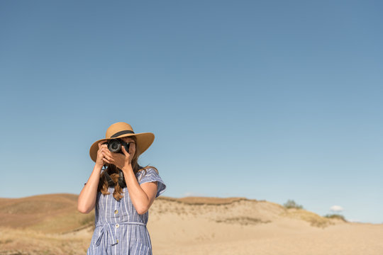 adult woman in straw hat and dress with camera taking photo on sand dune of beach in a sunny day