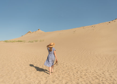 adult woman in casual dress and straw hat walking having good time on sand dune on hot summer day