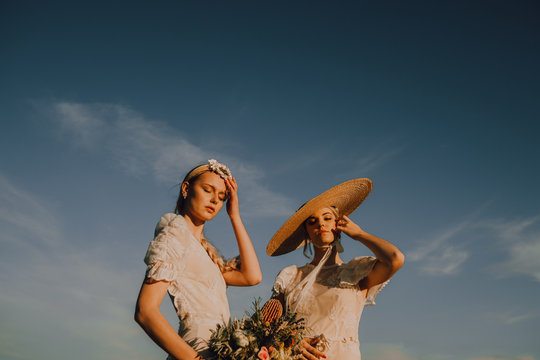 From below slim tanned women in wreath and straw hat with bunch of flowers on background of blue sky