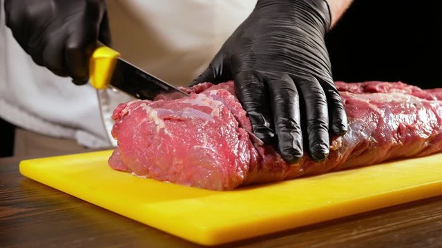 A professional butcher in black gloves slices a piece of raw meat for steak from entrecote with a sharp knife. The cook makes a preparation for cooking on the grill. Close-up.