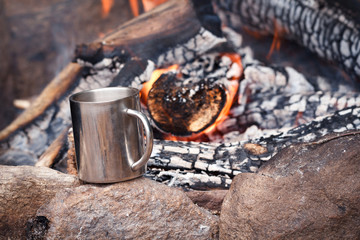 Stainless mug of hot drink standing on a stone by an outdoor campfire. Travel background. Campsite,...