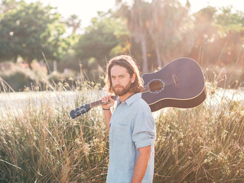Pensive bearded hipster man walking in jungle with guitar on shoulder looking at camera