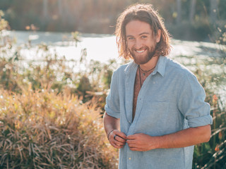 Young cheerful bearded hipster man unbuttoning blue shirt preparing to go swimming in river on sunny summer day
