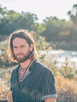 Young cheerful bearded hipster man near river looking at camera on sunny summer day