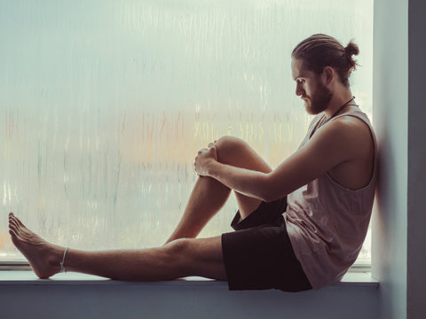 Side view of relaxed thoughtful male sitting in harmony on sill near against steamy window and looking down