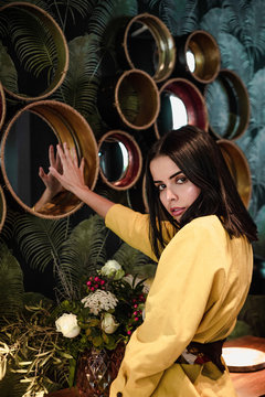 Pretty trendy young dark-haired female in fashionable outfit touching mirror by hand while looking at camera near flowers and plants indoors