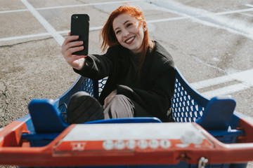 Attractive young woman with red hair smiling and taking selfie sitting in blue shopping cart with red handle