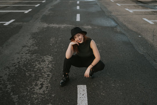 From above attractive young woman with red hair in black hat smiling and sitting on road with white stripes markup