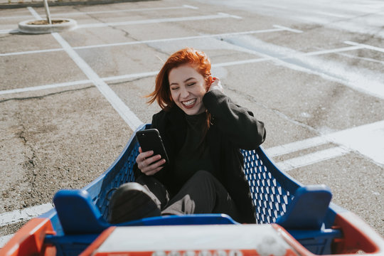 Smiling woman with smartphone in shopping trolley in parking lot