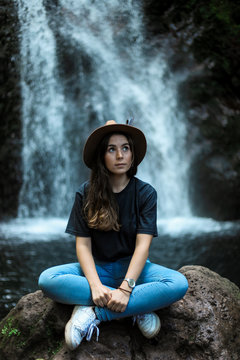 Cheerful female traveler in hat smiling and looking at camera while sitting on wet boulder near waterfall