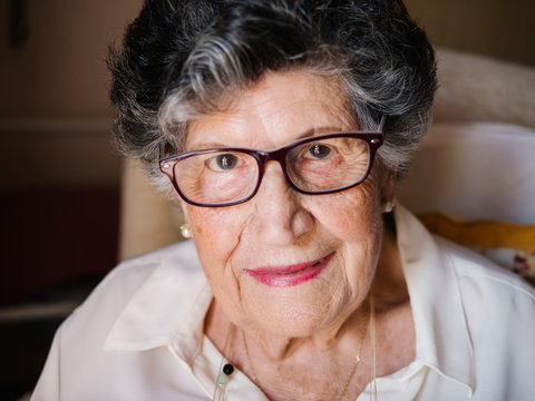 Smiling aged curly gray haired female in glasses with pink lips looking at camera in apartment