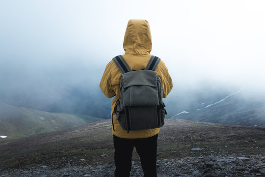 Back view of guy with backpack standing on hillside against thick fog during trip in nature