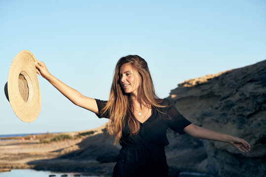 Young long haired smiling female model looking away holding hat in air on beach