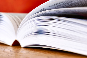Open text book with visible pages on wooden desk against a red background. Back to school, education or study concept. Closeup side view shot with selective focus.
