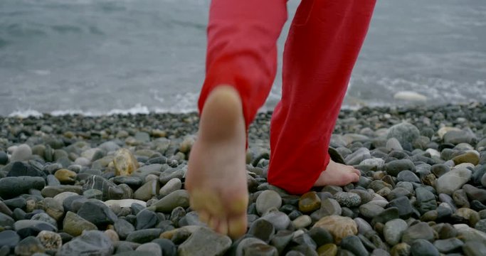 barefooted woman is stepping over large pebbles on beach, moving to sea water line