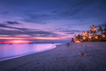 Sandy beach with palm trees at colorful sunset in summer.  Tropical landscape with sea shore, blurred water, palms, boats and yachts in ocean, purple sky with clouds at night. Travel in exotic Africa