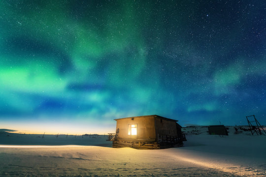 Aurora borealis over old small house with yellow light from the window. Northern lights in Teriberka, Russia.  Blue sky with stars and green polar lights. Night winter landscape with aurora, building