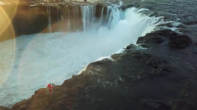 Aerial, drone shot, panning around a woman, taking pictures of the Godafoss, the waterfall of the gods, at the Skjalfandafljot river, Bardardalur district, at sunset, on a sunny evening, in Iceland