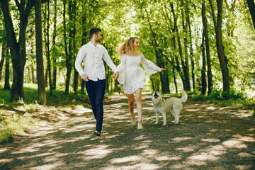 A beautiful and gentle girl with light hair and a white dress is walking in a sunny summer forest with her handsome guy in a white shirt and dark pants and they playing with cute gray dog