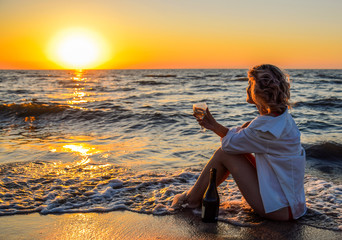 Girl with a glass and a bottle of champagne sits on seashore. © eleonimages