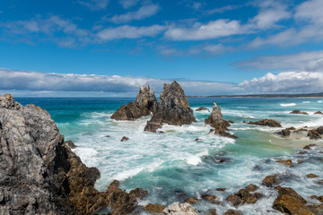 Mountain-like rocky formations cover the beach as the waves of water come crashing in on a cloudy day in Camel Rock Australia