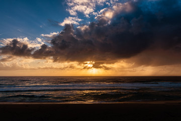 Naklejka na ściany i meble Strong sunrise dominates the dark and mysterious clouds that limit the sun's coverage to shimmers across the ocean, on Terrace Beach in NSW Australia