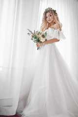 The bride's fees at the hotel. A beautiful blonde is standing at the window in a wedding dress. Photo with a bouquet.