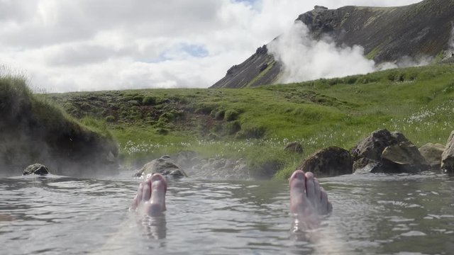 4K handheld close-up shot in daylight of two feet sticking out of the steaming water in a geothermal river with a background view of the mountains of Reykjadalur in Iceland.
