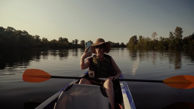Woman Taking Selfie On A Kayak Boat On Evening.Girl Making Self Photo And Answer To Video Call.Selfie On Mobile Phone In Beautiful Place On Tranquil Water Pond At Sunset Time.