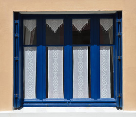 Sonniger Blick auf ein Fenster mit blauen Fensterläden und einer Hauswand, in dem eine gehäkelte,...