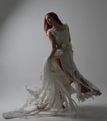 full length portrait of red haired girl wearing torn and tattered wedding dress. Standing pose against a studio background with contrasty shadow lighting.