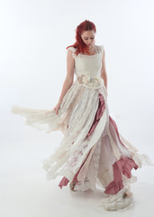 full length portrait of red haired girl wearing torn and tattered wedding dress. Standing pose against a  white studio background.