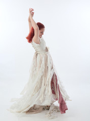 Fototapeta na wymiar full length portrait of red haired girl wearing torn and tattered wedding dress. Standing pose against a white studio background.