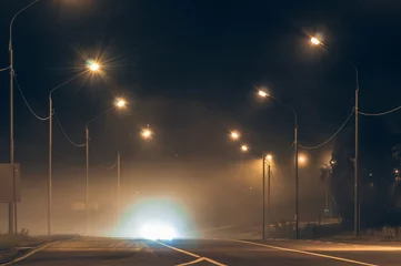 Tuinposter foggy night road,dim yellow light of street lamps. headlights of an approaching car.asphalt road. city night landscape © Екатерина Белоусова