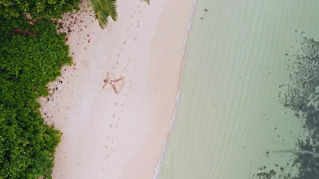 4k aerial vertical move up footage from girl doing sand angel on white tropical beach. Calm ocean waves and palm trees around