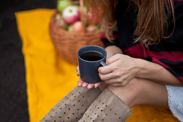 Outdoors autumn picnic. Young girl sitting on a yellow blanket and holding mug of hot coffee in a...