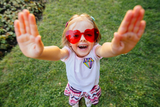 Little baby girl in red eyeglasses heart form with raised arms to give high five and looking at camera, toothy smile. close up portrait. happiness, love, emotion and feeling