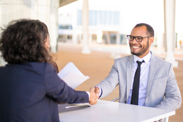 Successful business people meeting outside and closing deal. Happy business man and woman sitting at table, talking and shaking hands. Deal or success concept