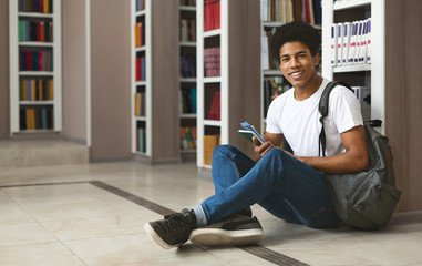 Cheerful afro student sitting on library floor next to bookshelves