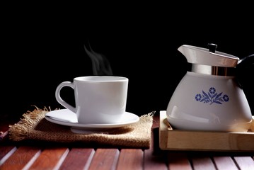 Hot tea or coffee in vintage white ceramic teacup with steam and teapot on wooden tabletop in black background, hot drink concept
