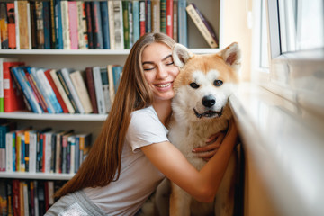 Fototapeta na wymiar Young woman sitting near window in front shelf of books, home library and hugs her dog. Beautiful young girl in library with her cute funny pet.