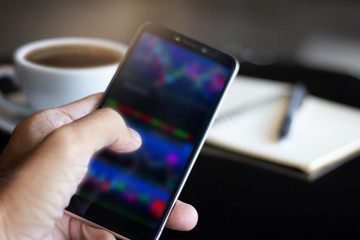 Closeup of hand businessman using smartphone working with coffee and notebook on black table. Concept you can do business on mobile and trading stocks anywhere, blur set index of stocks on screen