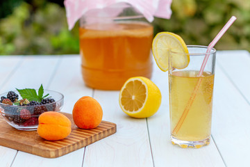 Glass jar with kombucha, a poured glass with kombucha with a slice of lemon, fruits on a wooden board on a light background in the summer garden.