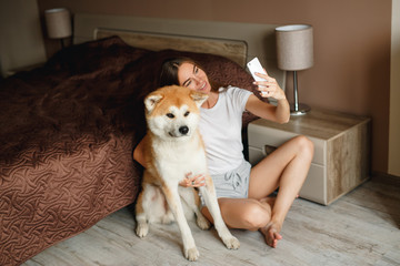 Beautiful young girl in casual clothes is making a selfie with her cute dog while sitting in bedroom, both smiling