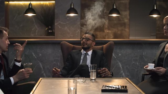 Dollying medium shot of male and female company executives in suits sitting in private hotel lounge with coffee and drinks, Middle Eastern businessman smoking cigar, and woman writing in organizer