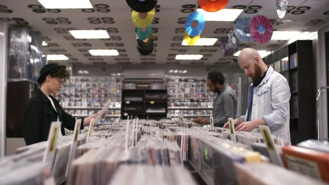 Trucking shot of multiethnic customers browsing through records in music shop, and stylish Caucasian woman in thirties, wearing bowler hat and glasses, choosing two vinyls in white covers and leaving