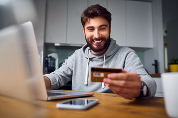 Portrait of smiling young man making online payments from home with credit card and laptop