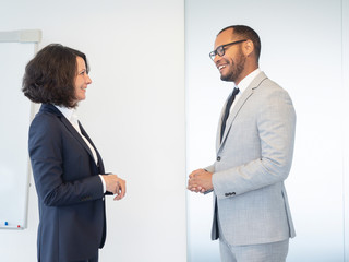 Two friendly confident professionals discussing business issues in conference room. Business man and woman standing indoors near white board and chatting. Business communication concept