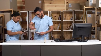 Full shot of two multiethnic post office clerks in blue uniform polo shirts returning to workplace, walking behind counter, sorting pile of envelopes into compartments then turning to work on computer